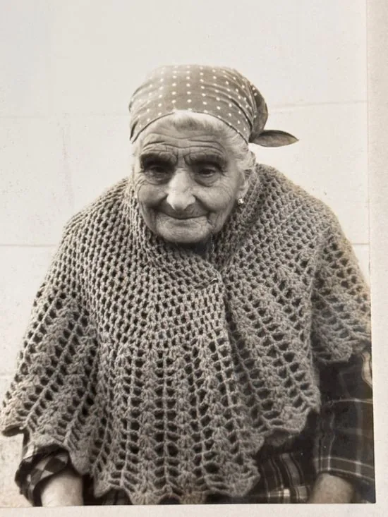 An elderly woman in a spotted headscarf and a thick crocheted shawl, smiling slightly.