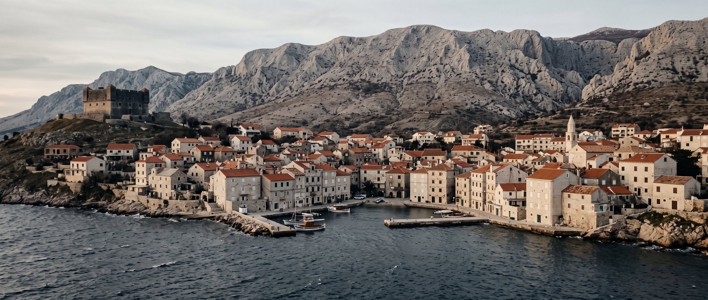 A stone town on the northern Adriatic beneath a bare karst mountain wall, a fortress on the hill above it and an almost empty harbour below.