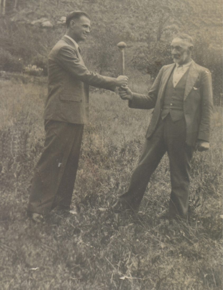 Two men shaking hands in open grassland, one young in a 1930s suit, one elderly with a white moustache and walking stick.