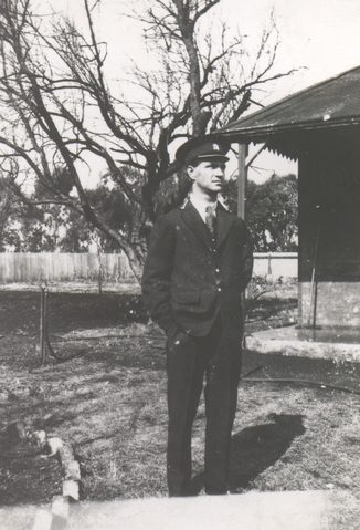 A young man in a dark uniform tunic and peaked cap standing in a yard by a bare tree.