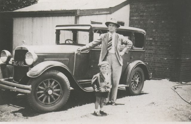 A man in a light suit and fedora leaning on a late-1920s sedan with a small boy beside him.